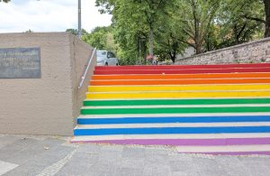 Zu sehen ist auf der rechten Seite eine Treppe, deren Stufen in Regenbogenfarben angemalt sind. Links daneben befindet sich eine Mauer und einer Gedenktafel mit der Aufschrift "Wo man Bücher verbrennt, verbrennt man auch am Ende Menschen" -Heinrich Heine.