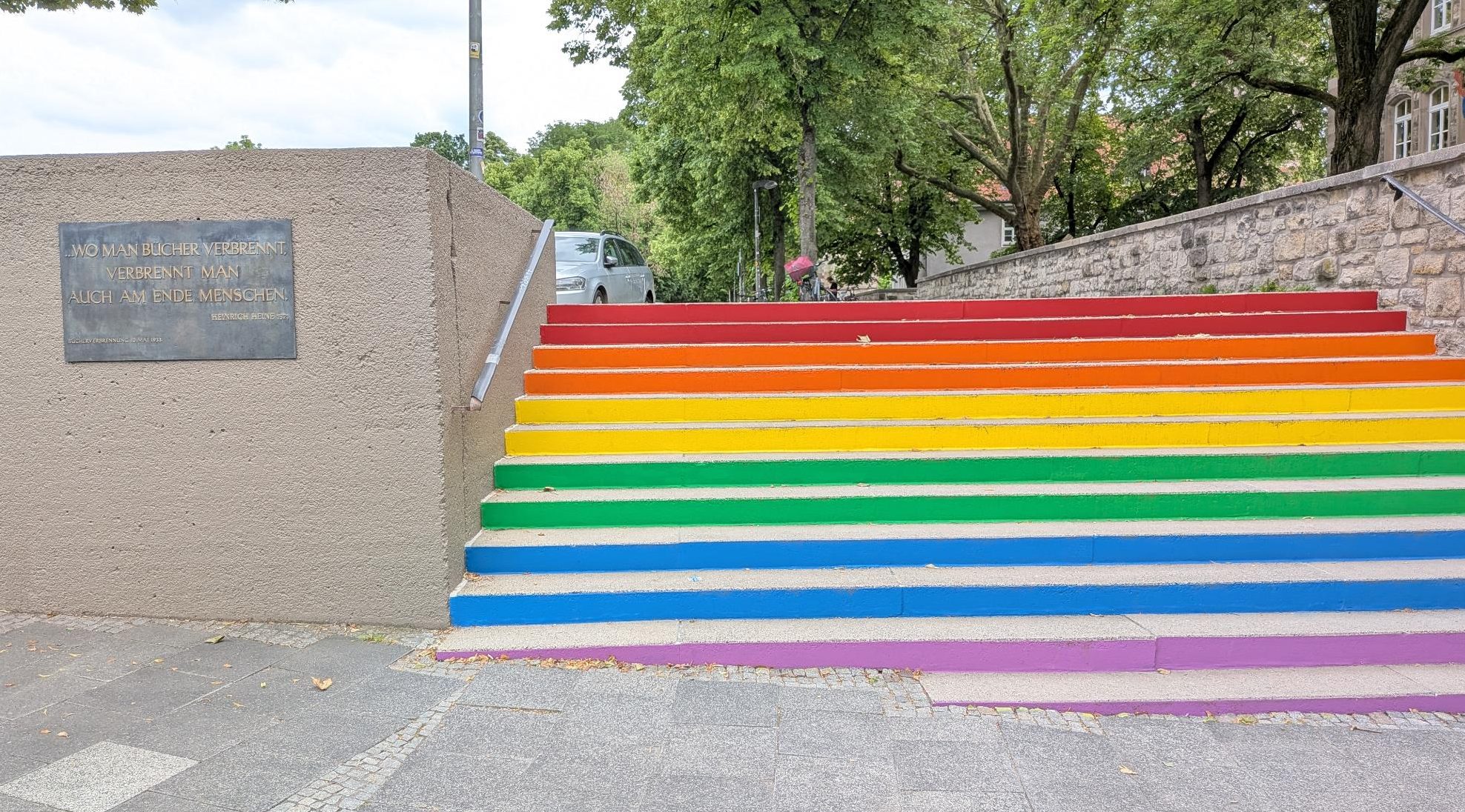 Zu sehen ist auf der rechten Seite eine Treppe, deren Stufen in Regenbogenfarben angemalt sind. Links daneben befindet sich eine Mauer und einer Gedenktafel mit der Aufschrift "Wo man Bücher verbrennt, verbrennt man auch am Ende Menschen" -Heinrich Heine.